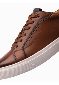 Sneakers en cuir brun avec une finition lisse, présentant une semelle en caoutchouc blanche, des accents texturés et des lacets bruns. Design minimaliste avec des perforations.