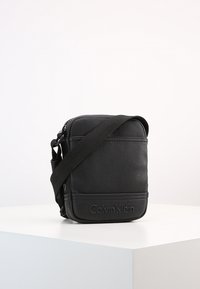 Borsa a tracolla nera in ecopelle testurizzata, chiusura con zip e cinturino regolabile. Presenta il logo Calvin Klein impresso sul davanti.