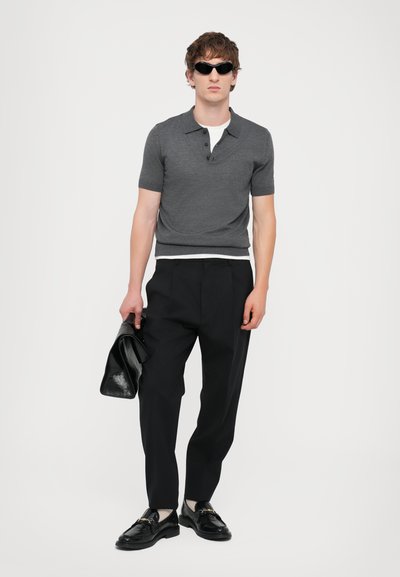 Grå kortærmet polo shirt, sorte bukser, sorte sko med hardware-detaljer. Modellen holder en sort jakke. Glat tekstur, skræddersyet pasform.