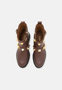 Botas de tobillo de cuero marrón con tachuelas doradas en forma de pirámide, un diseño de doble correa y una suela negra texturizada.
