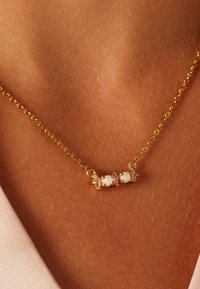 Gouden ketting met drie witte kralen afgewisseld met roze accenten, tegen een warme huidtint achtergrond.
