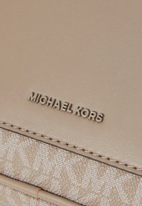Beige faux läderhandväska med texturerad tygpanel, med präglat mönster och metallisk logotyp. Inkluderar sömdekoration längs kanterna.