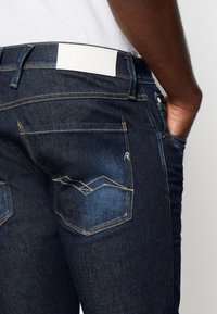 Baksidan av mörkblå denimjeans med vit "REPLAY"-etikett, ljusa sömmar och hand nedstoppad i framfickan.
