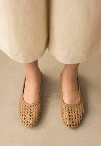 Flätade naturfiber lägenheter i beige med öppet vävdesign, rund tå och släta kanter, bärs med ett ljusfärgat, löst sittande plagg.