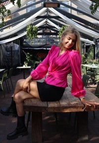 Satenasta bluza fuksije s napihnjenimi rokavi in pasom; kombinirana s črno mini krilom in črnimi gležnarji, sedi na lesenem mizi.