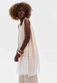 Beige mouwloos topje met een doorschijnende witte ruffle zoom, met een losse pasvorm en een gladde textuur, aangevuld met grote houten armbanden.