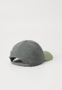 Căciulă din țesătură gri cu bor verde, având un buton superior cusut și o curea ajustabilă în spate. Textură netedă, design casual.
