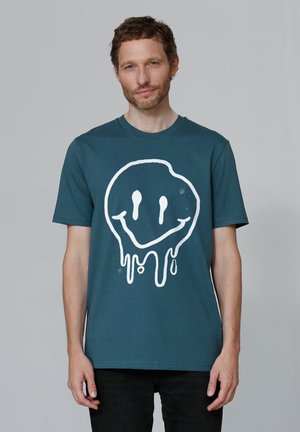 Turquoise katoenen T-shirt met een grote witte druipende smiley gezichtsafbeelding op de voorkant. Korte mouwen en een ronde hals.