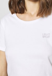 Vrouw in een eenvoudig wit T-shirt met ronde hals, met een klein "LIU JO SPORT" straslogo op de bovenste linkerborst.