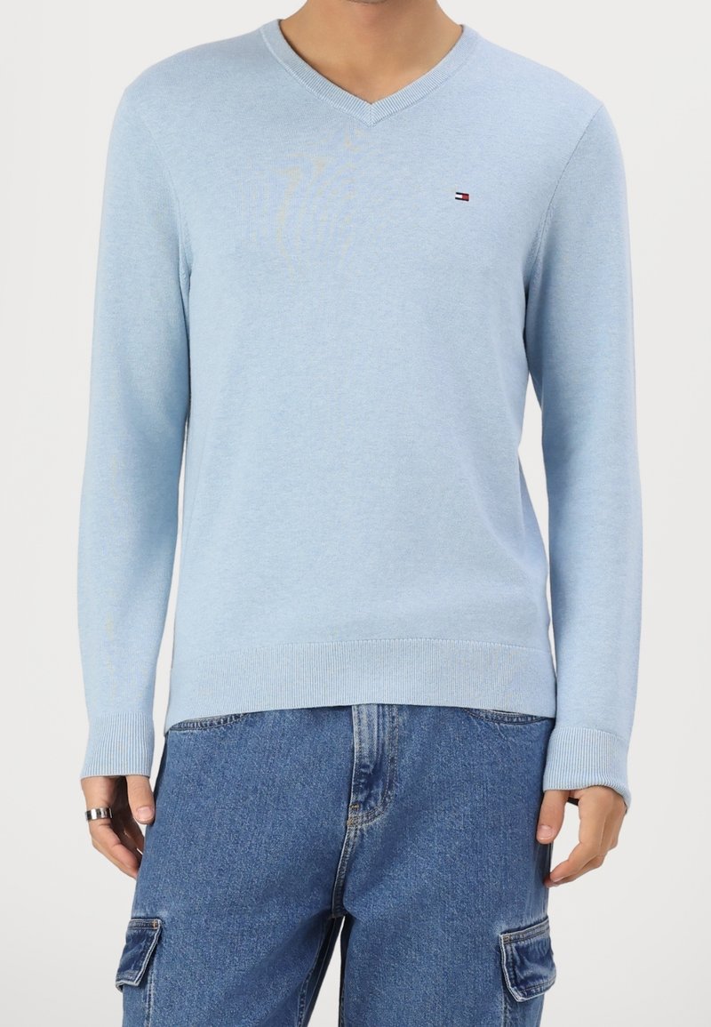 Homme portant un pull bleu clair à col en V avec un petit logo sur la poitrine et un jean cargo en denim bleu, debout devant un fond blanc.