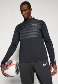 Musta spordiülikond polsterdatud tikitud rinnaosaga, millel on valged Nike'i ja Spursi logod. Mudel hoiab musta ja valge mustriga jalgpalli.