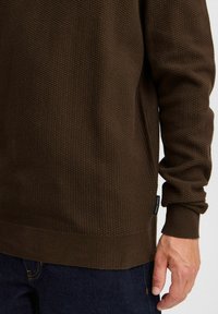 Brown textured sweatshirt with a honeycomb pattern, ribbed cuffs, and hem. Worn over dark jeans, showing a close-up of the sleeve and side.