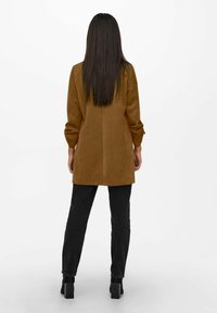 Hnědý suédé dlouhý kabát s volným střihem, dlouhými rukávy a centrálním švem na zadní straně, doplněný o černé slim-fit kalhoty a robustní podpatky.