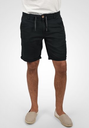 Uomo che indossa pantaloncini neri con orlo arrotolato, camicia bianca parzialmente visibile e scarpe beige slip-on, in piedi davanti a uno sfondo semplice.