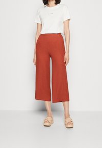 Femme portant un haut blanc à manches courtes et un pantalon ample taille haute couleur rouille, coupé au niveau des chevilles, avec des sandales compensées beige.