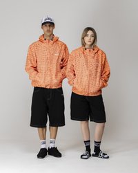 Orange gestreifte Jacken mit Kapuzen, schwarze Shorts und kontrastierende Schuhe. Die Jacken haben ein geometrisches Muster und Vordertaschen.