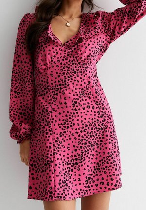 Mujer con un vestido rosa de manga larga con manchas negras de leopardo y un escote profundo con volantes, de pie contra una pared blanca.