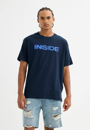Camiseta de algodón azul marino con la palabra "INSIDE" impresa en azul claro. Llevada con pantalones cortos de mezclilla claros y desgastados que presentan bordes deshilachados y grandes rasgaduras.