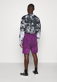 T-shirt graphique noir et blanc à manches longues, shorts violets unis et baskets noires avec semelles blanches. Les shorts ont deux poches arrière.