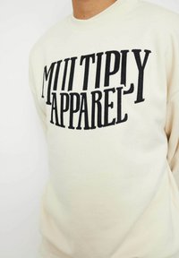 Sudadera crema con gran texto bordado en negro "MULTIPLY APPAREL". Diseño clásico de cuello redondo con una textura suave.