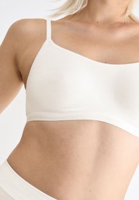 Vaaleanbeige saumaton bralette, jossa on säädettävät olkaimet. Pehmeä puuvillakangas, sileä pinta, eikä näkyvää metalliosaa tai koristeita.