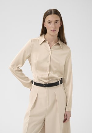 Žena s dlhými rovnými vlasmi, oblečená v béžovej saténovej košeli na gombíky zasunutej do širokých nohavíc rovnakej farby so čiernym opaskom, stojaca s jednou rukou na boku.