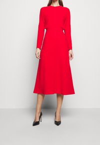 Vestido rojo de manga larga con corsé ajustado y falda acampanada, hecho de una tela suave. Tacones altos negros y puntiagudos completan el conjunto.