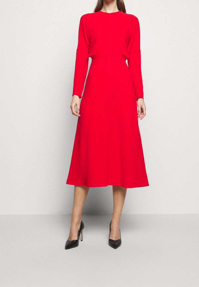 Vestido rojo de manga larga con corsé ajustado y falda acampanada, hecho de una tela suave. Tacones altos negros y puntiagudos completan el conjunto.