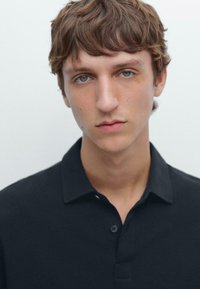 Svart polo skjorta med krage, med en knapp vid halsen. Modellen har kort, vågigt brunt hår och ljusblågröna ögon mot en neutral bakgrund.