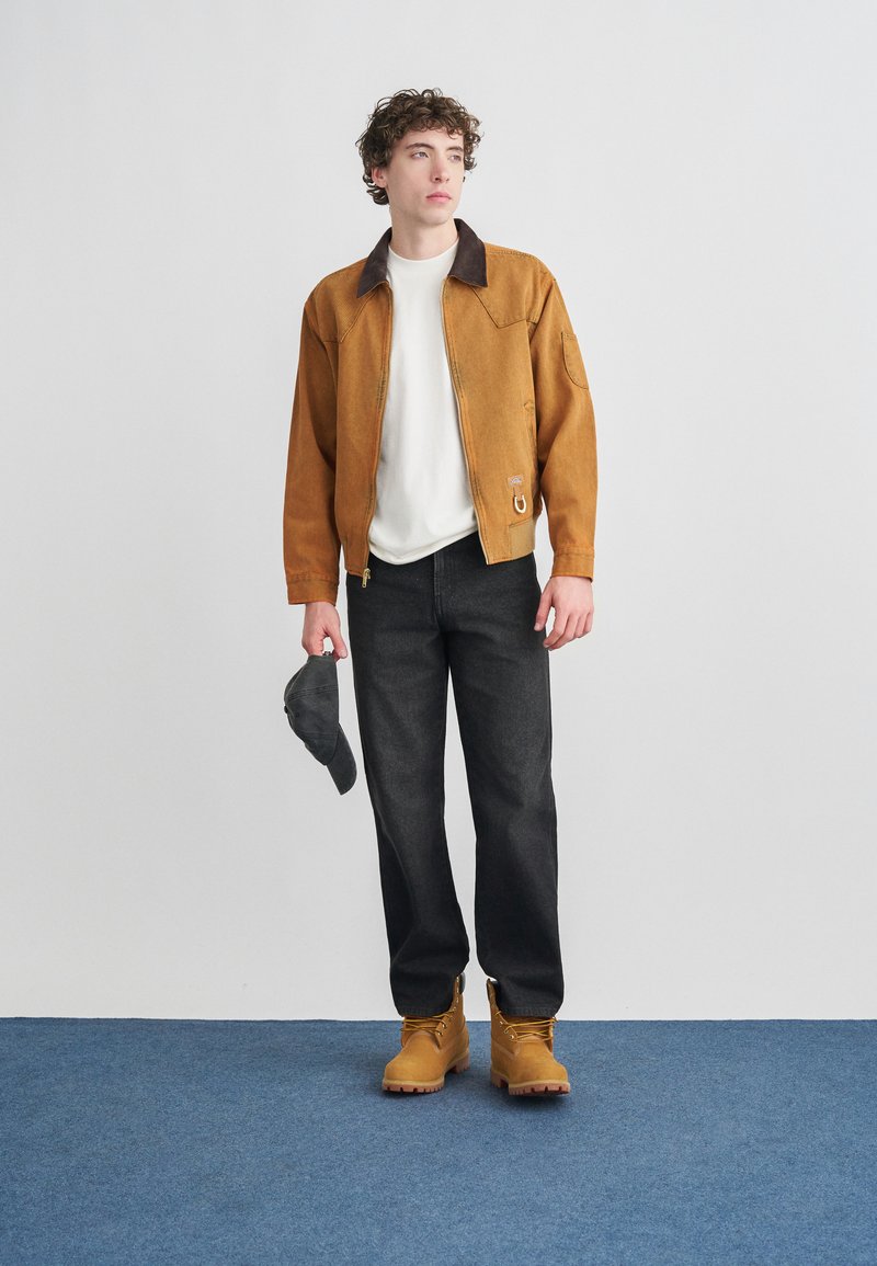 Brown jacket with a dark collar, white shirt, black jeans, and tan boots. The person holds a gray cap, standing on a blue carpet.