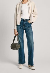 Ljusbeige bomberjacka, vit topp, vida blå jeans, off-white sneakers och en mörkgrön handväska som hålls i handen.