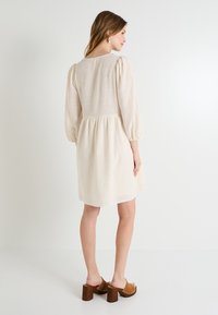 Vestido beige claro con cintura fruncida, mangas abullonadas y una textura suave, combinado con zuecos marrones de tacón con detalles dorados.