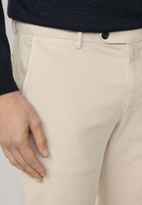 Beżowe spodnie z tkaniny bawełnianej o gładkiej powierzchni, z bocznymi kieszeniami i pojedynczym czarnym guzikiem zapięcia w pasie.