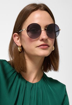 Donna con capelli castani fino alle spalle indossa grandi occhiali da sole rotondi scuri, orecchini a cerchio dorati e una maglietta verde plissettata su sfondo chiaro.