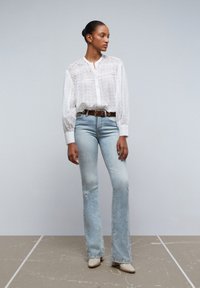 Blusa bianca in cotone con occhielli, con bottoni frontali, colletto alto e maniche lunghe; jeans flare azzurri con dettagli strappati; mule beige con tacco.