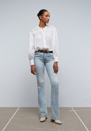 Blusa blanca de ojales con botones en el frente, cuello alto y mangas largas; vaqueros acampanados de color azul claro con un detalle desgastado; mule de tacón en color beige.