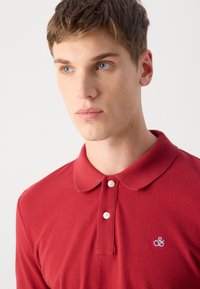 Rotes Polohemd aus glattem Stoff mit einer Knopfleiste mit zwei Knöpfen und einem blauen gestickten Logo auf der linken Brust, mit einem klassischen Kragen.