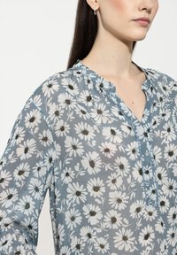 Blumenbluse in Hellblau, mit weißen Gänseblümchen und dunklen Mittelpunkten. V-Ausschnitt-Design mit Knopfleiste und aus weichem, leichtem Stoff.