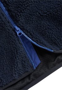 Marineblaue Fleecejacke mit einem blauen Reißverschluss, verfügt über eine strukturierte Oberfläche und ein schwarzes, perforiertes Innenteil. Der Reißverschluss ist teilweise geöffnet.