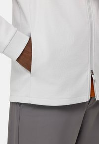 Chaqueta blanca de cremallera con tela texturizada y puños acanalados, combinada con pantalones grises. La chaqueta tiene bolsillos laterales.