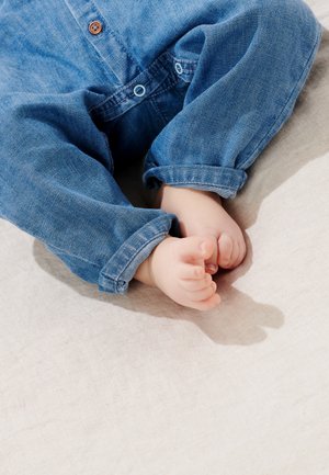 Denimblå opkørte ærmer og bløde beige knælange bukser, med små bare fødder hvilende på en lyst farvet stofoverflade.