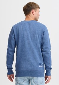 Blue sweatshirt with long sleeves, textured fabric, crew neck, and embroidered logo on the lower back. Paired with light denim jeans.
