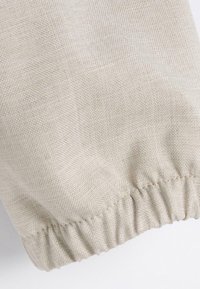 Lichtbeige stof met een gestructureerde weving en een geplooide elastische rand. Het materiaal lijkt zacht, met een subtiele glans en geen zichtbare patronen.