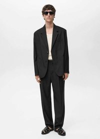 Completo nero con giacca e pantaloni a vestibilità ampia, abbinato a una camicia beige chiaro e sandali neri. Il modello indossa occhiali da sole scuri.
