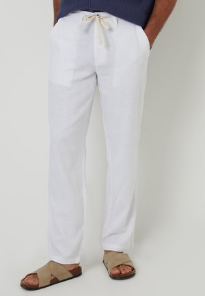 Mann trägt weiße Leinenhose mit Kordelzug und Taschen sowie beige Sandalen mit gekreuzten Riemen, steht mit den Händen in den Taschen.