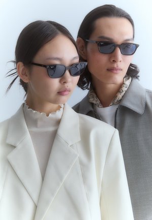 Ochelari de soare negri cu formă de ochi de pisică, o bluză albă cu volane și o jachetă gri cu un model, cu o cămașă deschisă și o eșarfă vizibile în fundal.