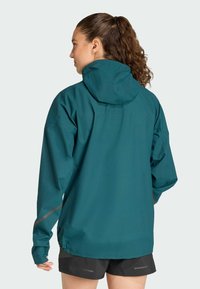 Teal waterproof jacket with a hood, loose fit, reflective accents on sleeves, and a slightly longer back. Thin fabric, minimal seams.