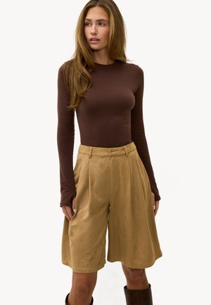 Femme portant un haut marron ajusté à manches longues et un short large à taille haute de couleur beige arrivant aux genoux, debout devant un fond blanc.