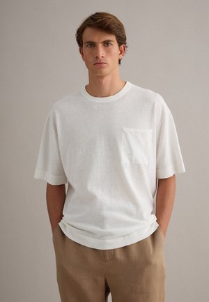 Junger Mann mit braunen Haaren, der ein weißes kurzärmeliges Hemd mit Brusttasche und beige Hosen trägt, vor einem schlichten hellen Hintergrund stehend.