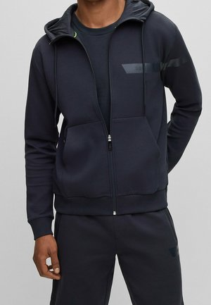Marinblå zip-up hoodie med justerbar huva, framfickor och diskreta logotypdetaljer. Tillverkad av mjukt, smidigt tyg med en figurnära design.